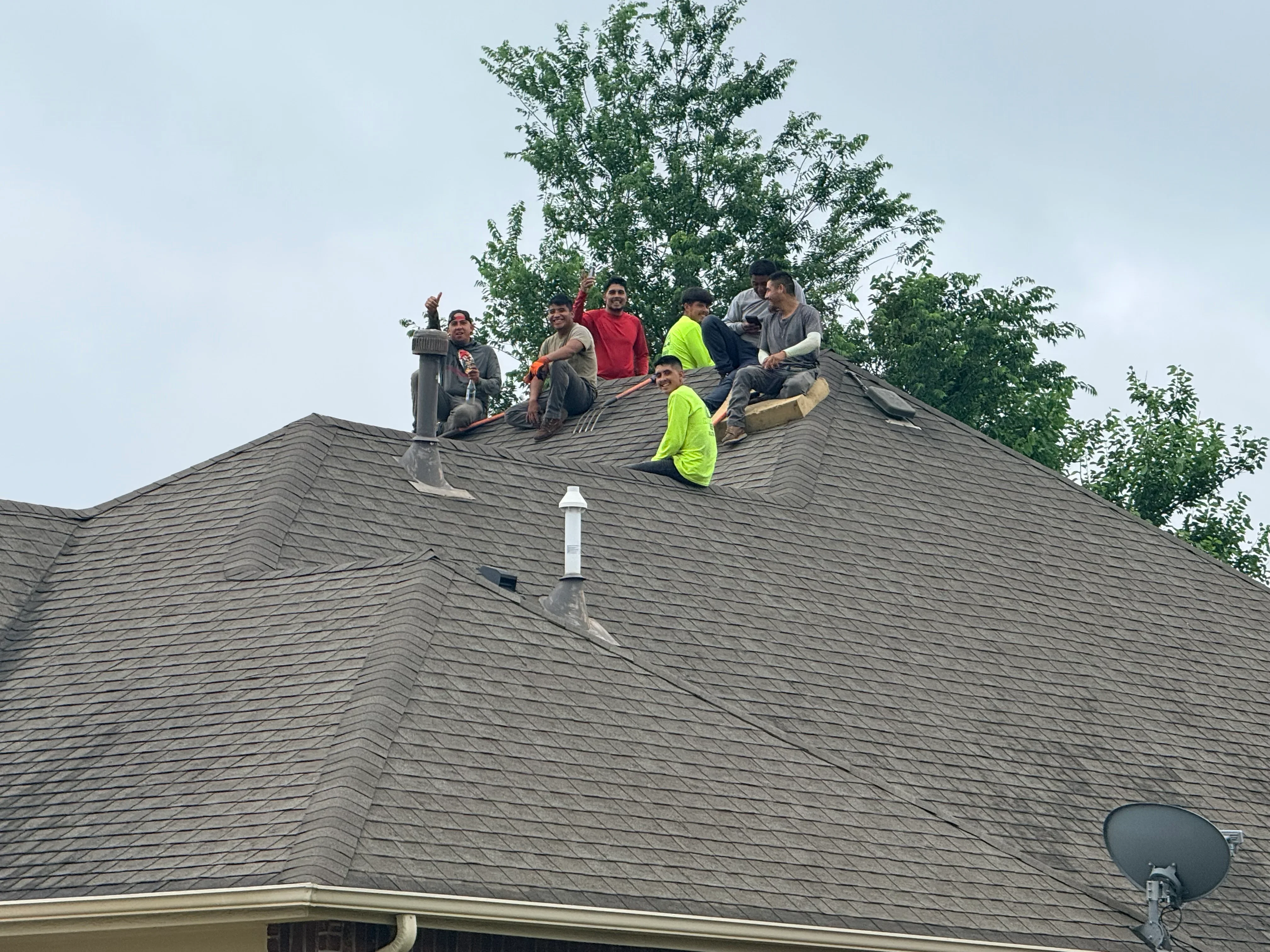 Crew installing gray architectural shingles on multi-slope residential roof with chimney flashing