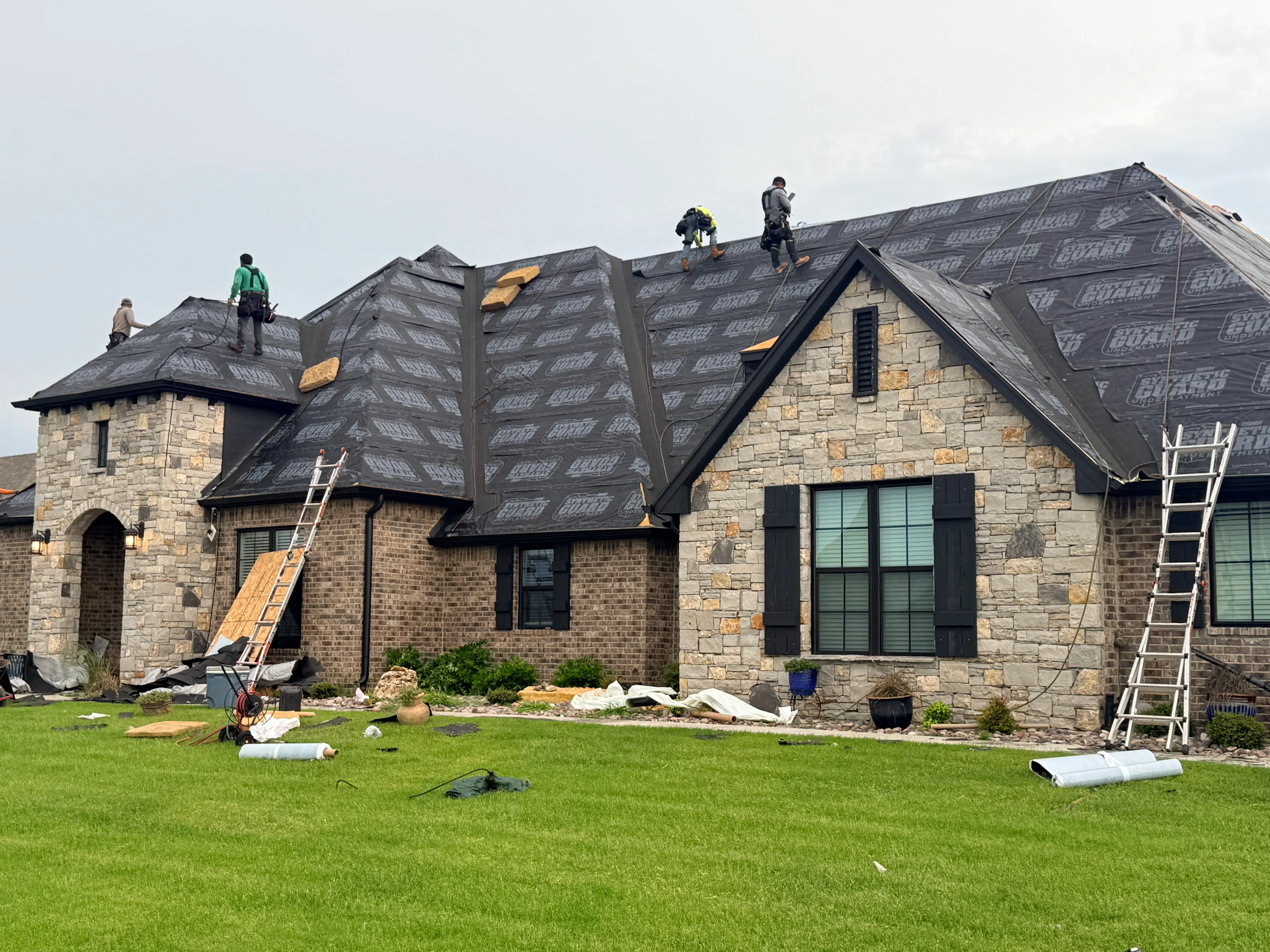 Roofers installing architectural shingles on multi-slope stone home with turret and gabled sections