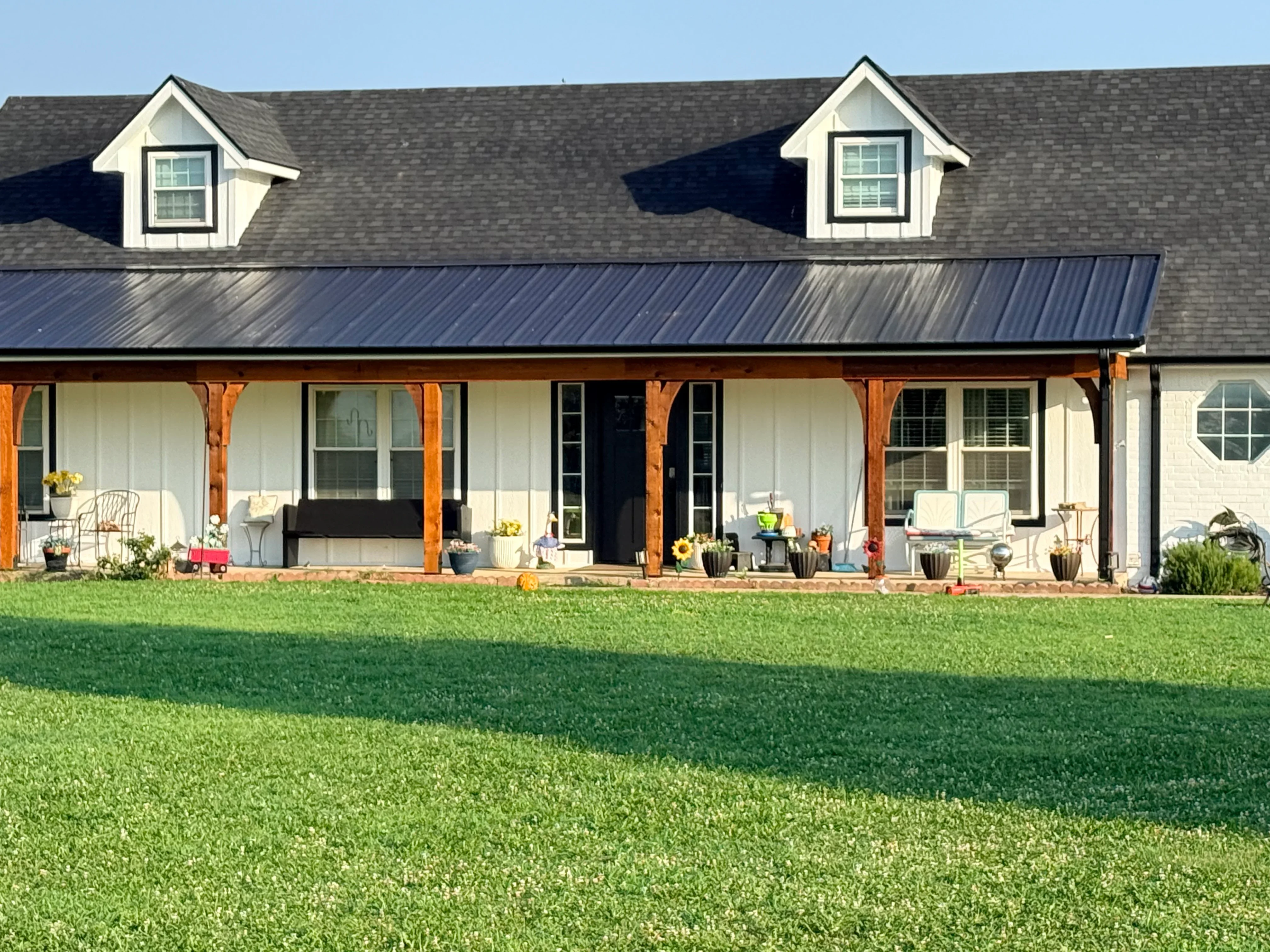 Standing seam metal roof with gray shingles, white dormers, and timber porch supports on farmhouse-style home
