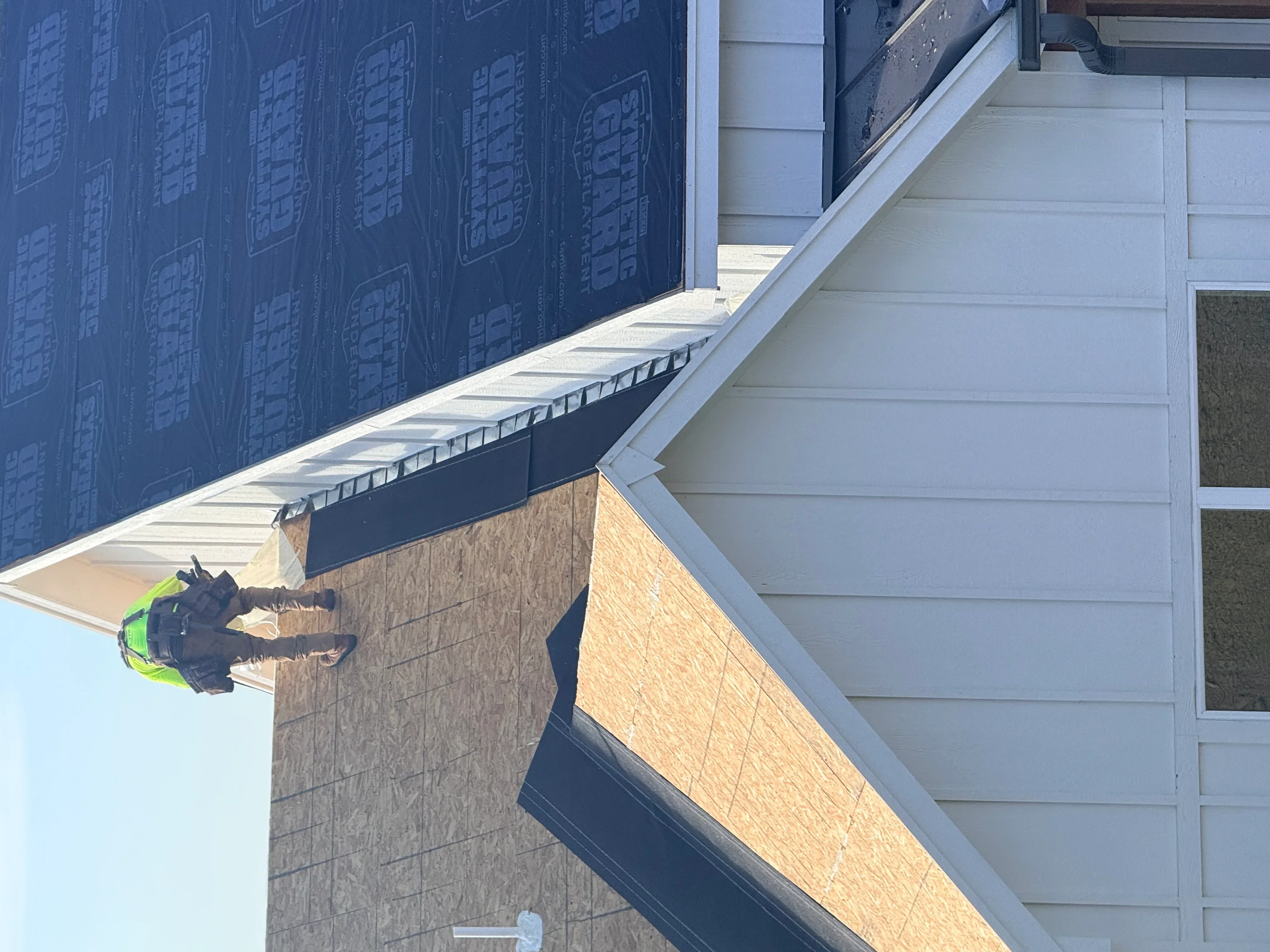 Roofer installing blue synthetic underlayment and white metal drip edge on residential roof with OSB sheathing